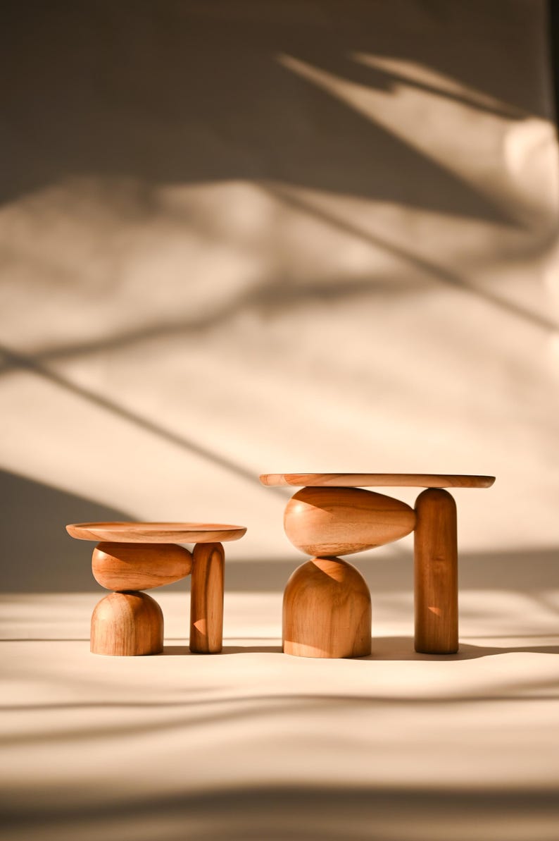 May include: Two wooden display stands of varying heights, each featuring a flat circular top supported by a base composed of a smooth, rounded dome-shaped piece, an oval-shaped element, and a single vertical cylindrical pillar.