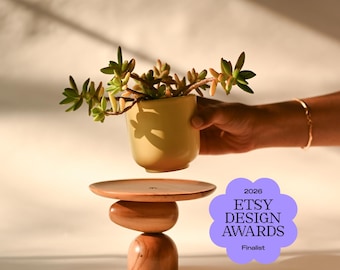 May include: A light yellow ceramic planter containing a green succulent, held above a wooden pedestal stand with a flat circular top and a stacked base. A purple flower-shaped badge displays the text 2026 ETSY DESIGN AWARDS Finalist.