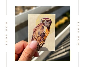 Puede incluir: Una pequeña ilustración pintada a mano de un búho con plumas marrones, naranjas y moradas. El búho mira directamente al espectador con grandes ojos oscuros. La ilustración está en una tarjeta blanca con un halo amarillo alrededor del búho.