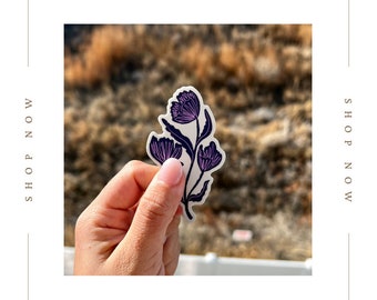 Puede incluir: Una pegatina con un diseño floral morado y negro con tres flores en un tallo. La pegatina se sostiene en una mano contra un fondo borroso.