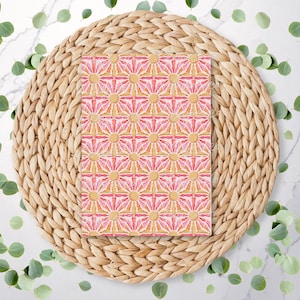 May include: A rectangular pattern featuring a repeating design of pink and yellow sunbursts. The pattern is set on a woven, circular placemat. Green leaves are scattered around the placemat on a white marble surface.