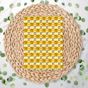 May include: A patterned paper with a repeating design of cherries and bows on a yellow background. The paper is placed on a round, woven, natural-colored placemat. Green leaves are scattered around the placemat.