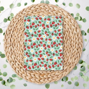Peut inclure: Un carnet à spirales avec un motif répété de fraises rouges, de marguerites blanches et de feuilles vertes sur un fond bleu clair. Le carnet est posé sur un dessous de plat rond en osier, avec des feuilles vertes éparpillées.