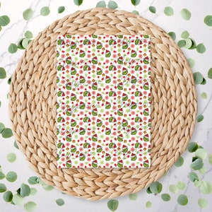 May include: A white rectangular item with a repeating pattern of a green creature wearing a Santa hat, red gifts, and red snowflakes. The item is on a woven, circular placemat, surrounded by green leaves.