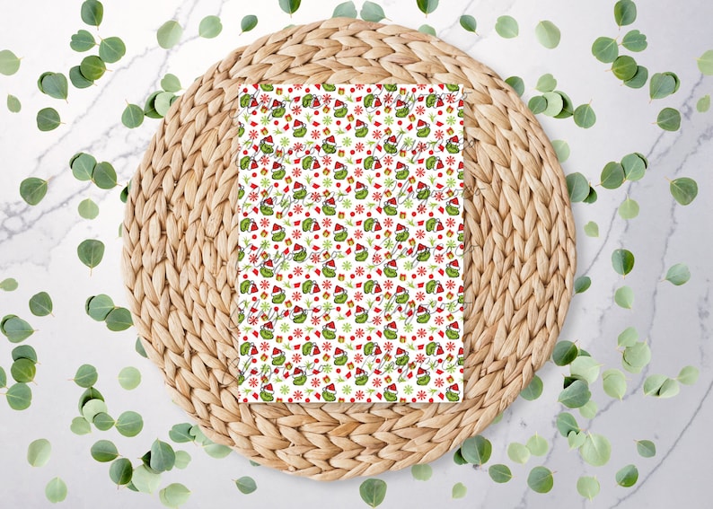May include: A white rectangular item with a repeating pattern of a green creature wearing a Santa hat, red gifts, and red snowflakes. The item is on a woven, circular placemat, surrounded by green leaves.