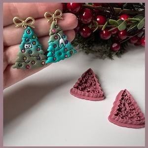May include: Two Christmas tree earrings with gold bows, one green and one teal, decorated with colourful ornaments. Two pink tree-shaped cookie cutters are also visible, along with red berries and greenery.