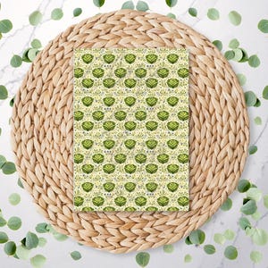 May include: A rectangular item with a repeating pattern of green floral designs on a light yellow background. The item is centered on a round, woven, light brown placemat, surrounded by scattered green leaves.