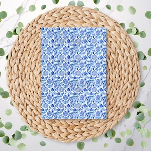 May include: A rectangular item with a blue and white floral pattern, featuring stylized flowers and ornaments, rests on a woven, circular placemat. Green leaves are scattered around the placemat on a white marble surface.