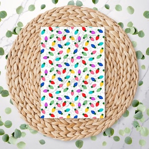 May include: A rectangular card with a white background features a pattern of colourful Christmas lights. The lights are red, green, blue, yellow, and pink. The card is on a woven, circular placemat, surrounded by green leaves.