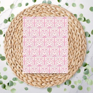 May include: A square card with a pink palm tree pattern on a white background. The card is centered on a round, woven, natural-colored placemat. Green leaves are scattered around the placemat on a white marble surface.
