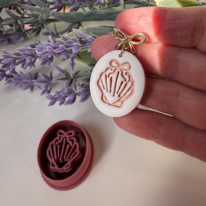 May include: A white oval pendant with a copper-colored seashell design and a gold bow-shaped bail. A matching seashell-shaped cookie cutter is also visible. The pendant is held in a hand, with lavender sprigs in the background.