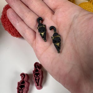 May include: Two small black cat figurines with gold-painted accents on the ears and chest, featuring curved tails. Below these, two matching pink plastic cookie cutters shaped like cats with detailed facial features are positioned on a white surface.