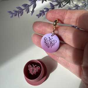 May include: A circular lavender clay charm featuring an embossed bee design attached to a gold-toned metal jump ring, accompanied by a matching circular pink plastic clay cutter with a raised bee-shaped stamp in the centre of the base.
