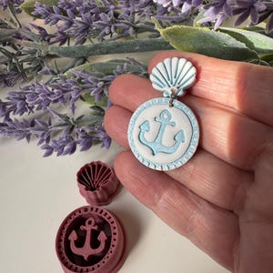 May include: A handmade nautical-themed earring with a white and light blue color scheme. The earring features a seashell-shaped top and a circular pendant with an anchor design. Two polymer clay cutters, one seashell and one anchor, are also visible.