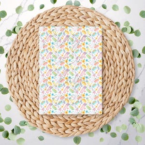 May include: A notebook with a repeating watercolour pattern of beauty products, including lipsticks, nail polish, and perfume bottles in pastel colours. The notebook is on a woven placemat with eucalyptus leaves scattered around.