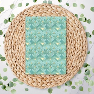 May include: A rectangular paper with a geometric pattern in shades of teal and gold, resting on a woven, circular placemat. Green leaves are scattered around the placemat on a white surface.