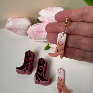 May include: A gold charm with a small, pink and white cowboy boot pendant. The boot has a heart design and a gradient effect. Also, there are three pink cowboy boot-shaped cookie cutters.
