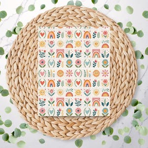 May include: A square notebook featuring a repeating pattern of colourful flowers, hearts, rainbows, and suns. The notebook is placed on a woven, circular placemat. Eucalyptus leaves are scattered around on a white marble surface.