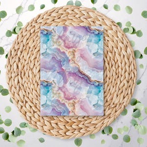 May include: A rectangular item with a colorful agate pattern in shades of blue, pink, and purple, resting on a woven, circular placemat. Green leaves are scattered around the placemat on a white marble surface.