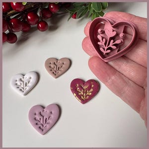 May include: Heart-shaped clay ornaments in white, pink, and burgundy, each with a floral design. A pink heart-shaped cutter is held in a hand, with red berries in the background. The ornaments are likely handmade.