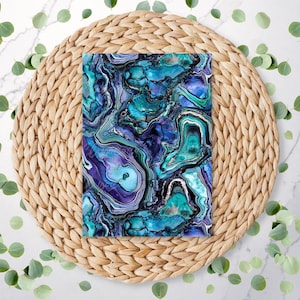 May include: A rectangular item with a swirling, abstract pattern in shades of blue, teal, and purple, resembling agate or marble. It rests on a round, woven, light brown placemat, surrounded by green leaves.
