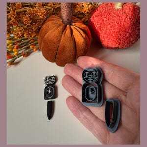 May include: Black cat-shaped cookie cutters with a clock design. The cutters are held in a hand, with a pumpkin and autumn foliage in the background. The cutters are for making Halloween or autumn-themed biscuits.