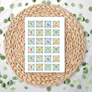 May include: A white card with a grid of watercolour illustrations of vegetables and plants. Each square features a different design, including carrots, tomatoes, aubergines, pumpkins, and broccoli, set against a blue and white tile pattern. The card rests on a woven, circular placemat.