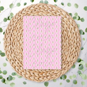 May include: A pink and white patterned paper with a floral design, set on a woven wicker placemat. The paper features a repeating pattern of pink flowers and green stems on a pink and white gingham background. Green leaves are scattered around the placemat.