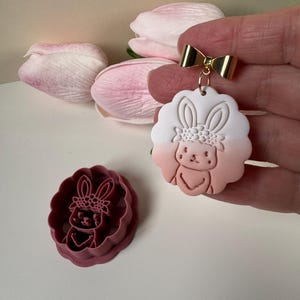 May include: A gold-coloured bow-shaped earring holder with a bunny-shaped charm. The charm is white and pink, with a bunny face and floral crown design. A matching bunny-shaped cookie cutter is also visible.