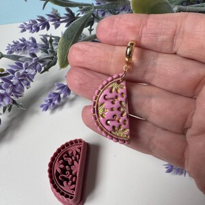 May include: A pink and gold-coloured half-circle pendant with a floral design. The pendant is attached to a gold-coloured hoop. A matching pink mould is next to the pendant. The background includes lavender and green foliage.