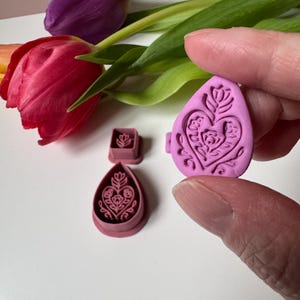 May include: Three pink polymer clay cutters in various shapes: a square, a teardrop, and an oval. Each cutter features a floral heart design. The background includes red and purple tulips with green leaves.