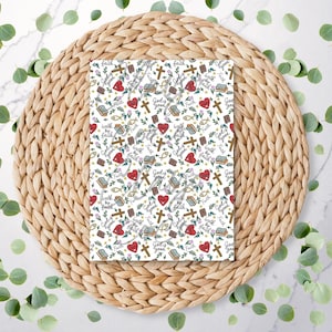 May include: A rectangular item with a white background, patterned with crosses, hearts, books, and fish symbols. The phrases "God is Good" and "Walk by Faith" are also visible. The item rests on a woven, circular placemat, with green leaves scattered around it.