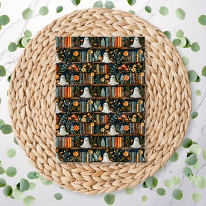 May include: A fabric with a repeating pattern of bookshelves, flowers, and white ghosts against a dark navy blue backdrop. The fabric is displayed on a woven, circular placemat, with green eucalyptus leaves scattered around it.