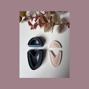 Peut inclure: Deux emporte-pièces en pâte polymère en forme de demi-cercle et de U, en bleu marine et rose clair. Le découpeur en forme de U a un motif géométrique. Une brindille de feuilles séchées est en arrière-plan.