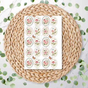 May include: A sheet of oval stickers with floral designs, including pink roses and butterflies. Each sticker has a light blue scalloped border. The stickers are on a white background, placed on a woven, circular placemat.