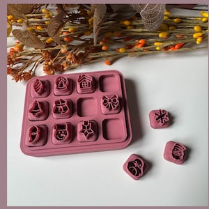 May include: A pink tray containing Christmas-themed cookie cutters, including a house, bow, and snowman. Several loose cutters are scattered nearby. The background features decorative foliage.