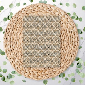 May include: A decorative placemat with a woven, natural fibre base. The placemat has a rectangular insert with a repeating, stylised floral pattern in beige and gold. Green eucalyptus leaves are scattered around the placemat on a white marble surface.