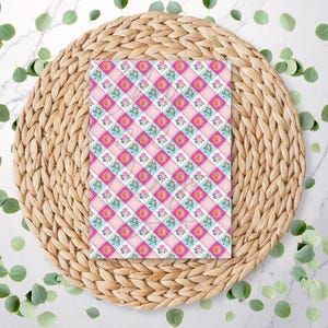 May include: A patterned paper with a pink, white, and blue floral design. The paper is placed on a round, woven, natural-colored placemat. Green leaves are scattered around the placemat on a white marble surface.
