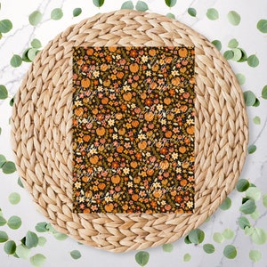 May include: A patterned paper with a black background, featuring a repeating design of orange pumpkins, yellow and orange flowers, and green leaves. The paper is placed on a woven, circular placemat, with scattered green eucalyptus leaves around it.