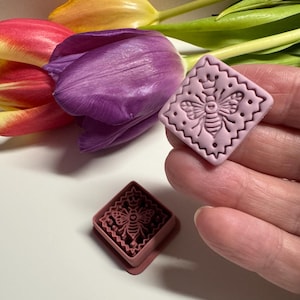 May include: Two pink square-shaped cookie cutters with a bee design. One cutter is held in a hand, and the other is on a white surface. The cutters have a decorative edge. Colorful tulips are in the background.