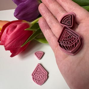 May include: A selection of pink polymer clay cutters in fan and teardrop shapes, featuring detailed patterns. The cutters are held in a hand. A red and purple tulip are in the background. The cutters are likely for crafting.