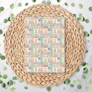 May include: A rectangular fabric with a patchwork design featuring pastel squares, each with a bunny, flowers, or Easter-themed elements. The fabric is placed on a woven placemat, surrounded by eucalyptus leaves.