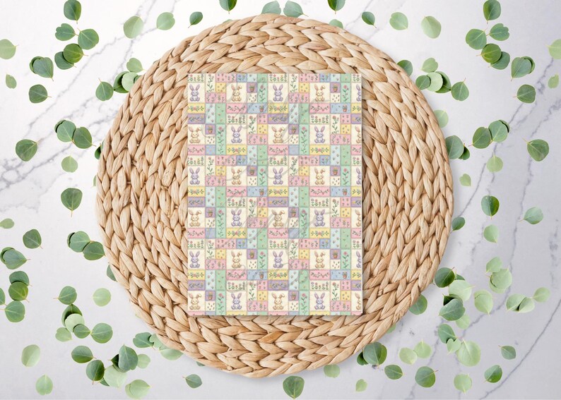 May include: A rectangular patterned fabric featuring a patchwork design with pastel squares. Each square contains a bunny, flowers, or other Easter-themed elements. The fabric is set on a woven placemat with eucalyptus leaves scattered around.