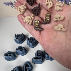 May include: A collection of small, moulded clay shapes featuring highland cows, horses, cowboy boots, and hats, alongside several blue plastic cookie cutters in matching shapes. The clay pieces are in shades of brown, tan, and cream with gold accents.