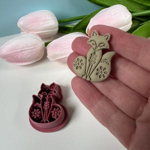 May include: A tan fox-shaped cookie cutter and a tan fox-shaped clay figure are displayed. The fox has floral details and is held in a hand. Pink tulips are in the background.