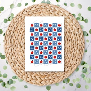 May include: A white card featuring a repeating pattern of red apples and white stars on a blue and white checkered background. The card is centered on a woven, circular placemat, with green leaves scattered around the edges.