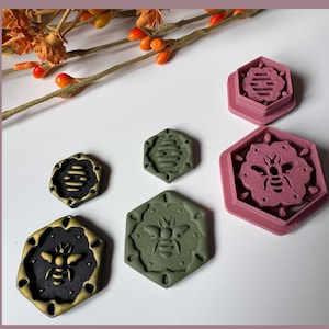 May include: A selection of hexagonal bee-themed stamps in black, gold, and pink. The stamps feature detailed bee and honeycomb designs. A sprig of orange berries is in the background. Measurements are in cm.