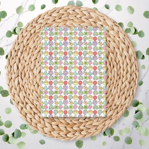 May include: A white card with a repeating pattern of colourful cartoon images. The images include a rainbow, a star, a heart, a cupcake, a balloon, and a teddy bear. The card is sitting on a woven straw placemat.