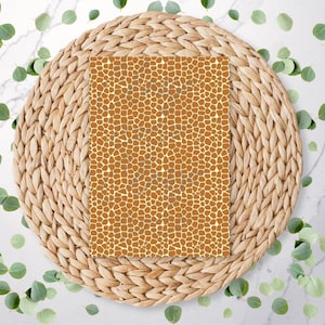 May include: A rectangular item with a giraffe print, featuring a pattern of brown spots on a cream background. The item is placed on a round, woven, light brown placemat, with green leaves scattered around.