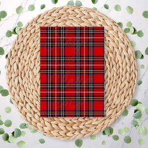 May include: A red plaid pattern with black, white, and yellow lines. The plaid is centered on a woven, circular placemat. Green leaves are scattered around the placemat on a white marble surface.